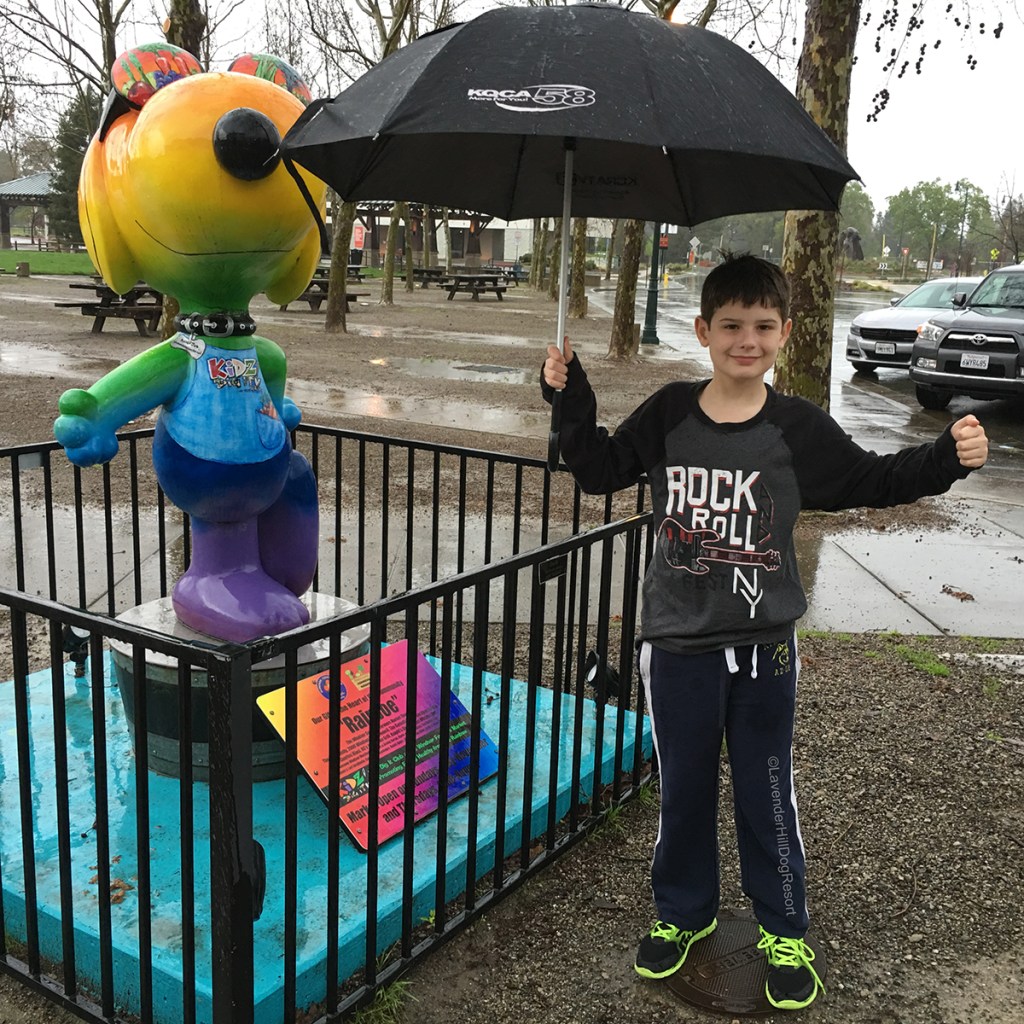 A boy outside standing next to a rainbow painted Snoopy statue.